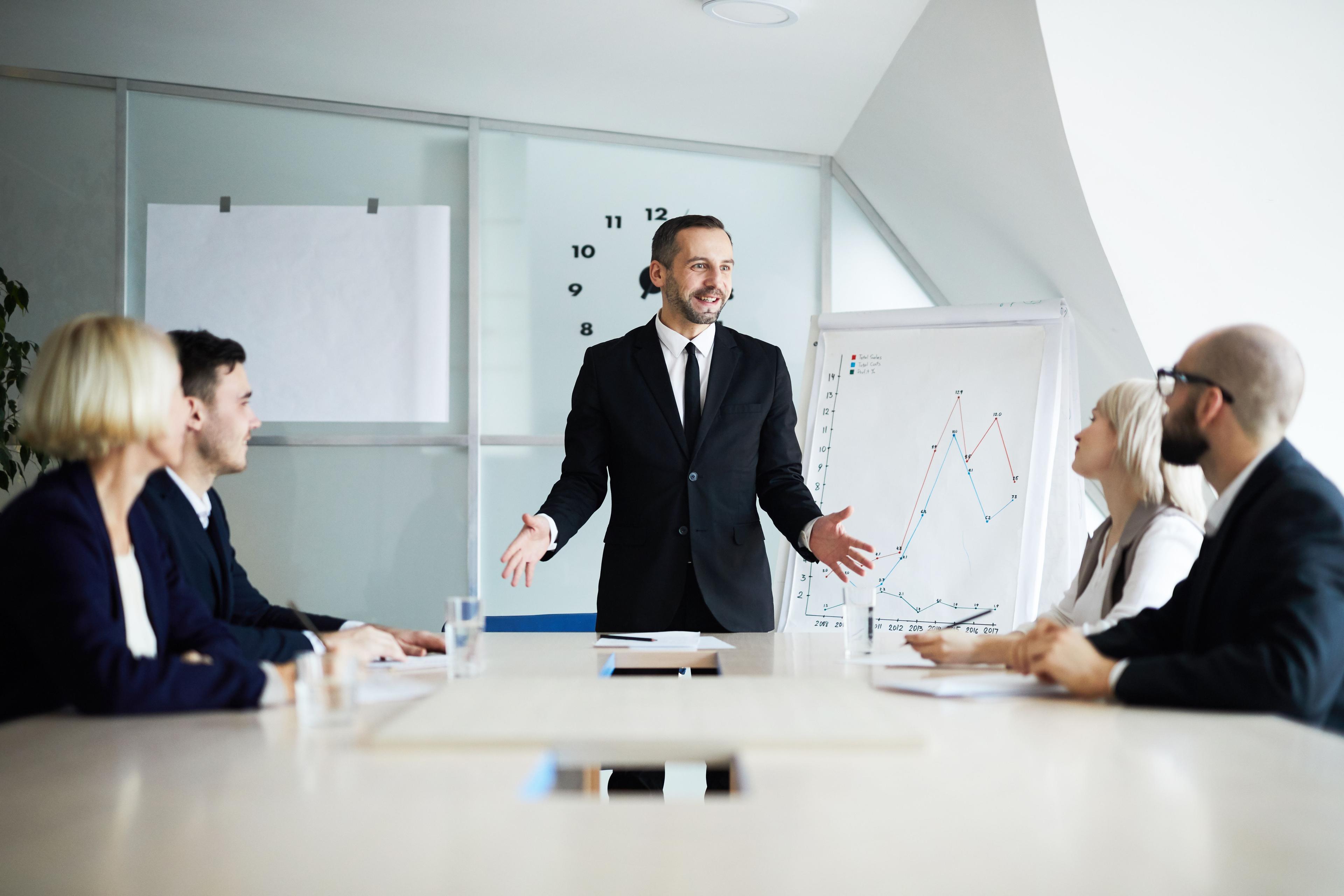 A confident business professional, dressed in a black suit, engaging a team in a lively discussion during a presentation, with a flip chart showing social media marketing metrics. Ideal for illustrating social media marketing jobs that require strong communication and leadership skills.