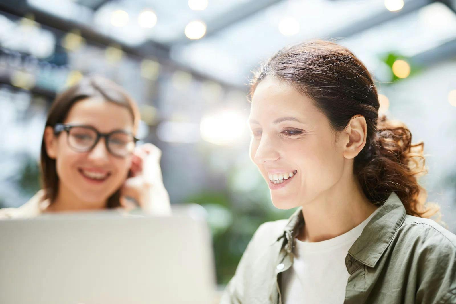 Two women collaborating on a laptop for real estate marketing, with one smiling and focused on the screen in a bright, modern workspace