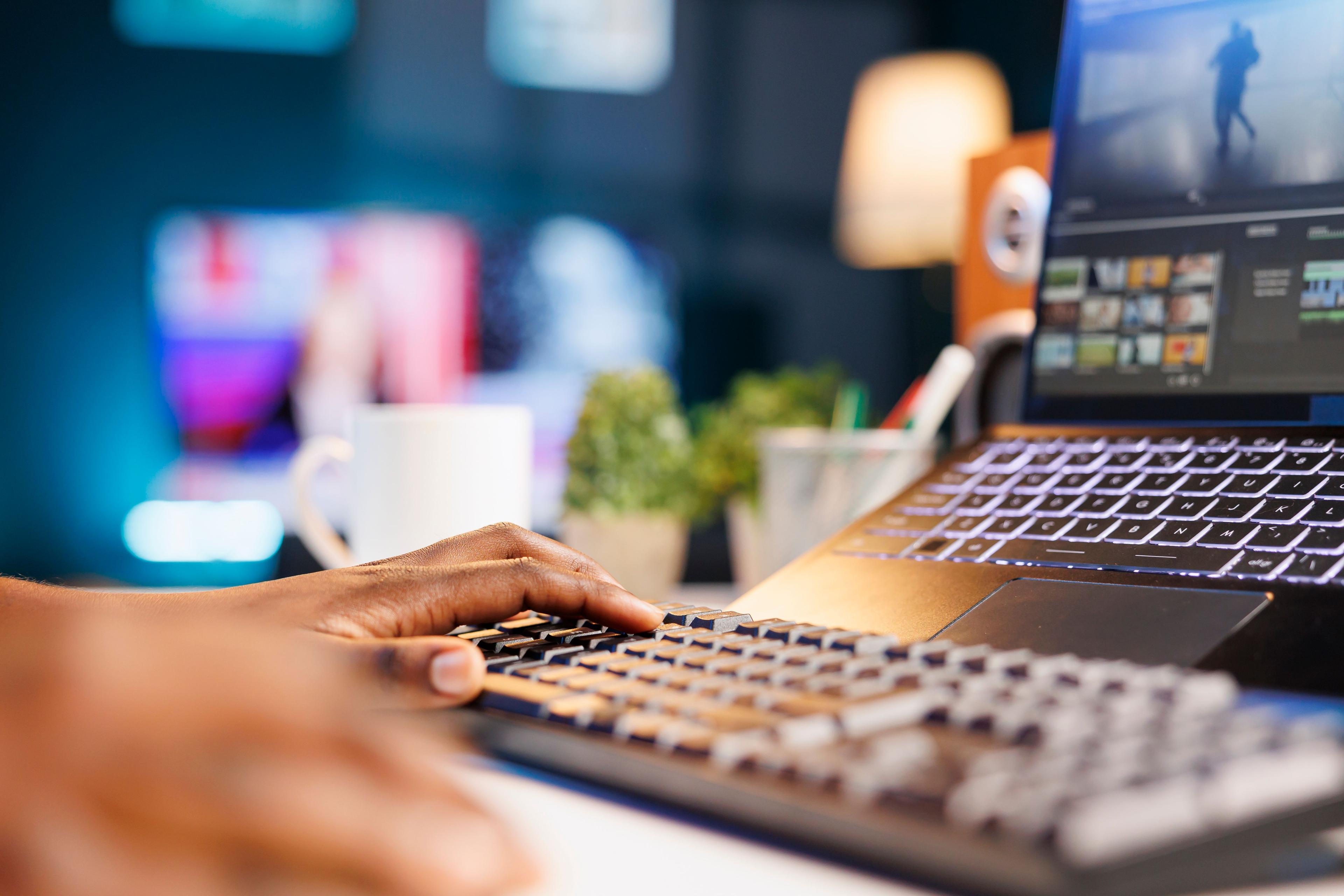 A close-up of a person's hands typing on a keyboard with a laptop screen displaying video editing software in a dimly lit room. The scene suggests a content creator working on a "social media marketing strategy."