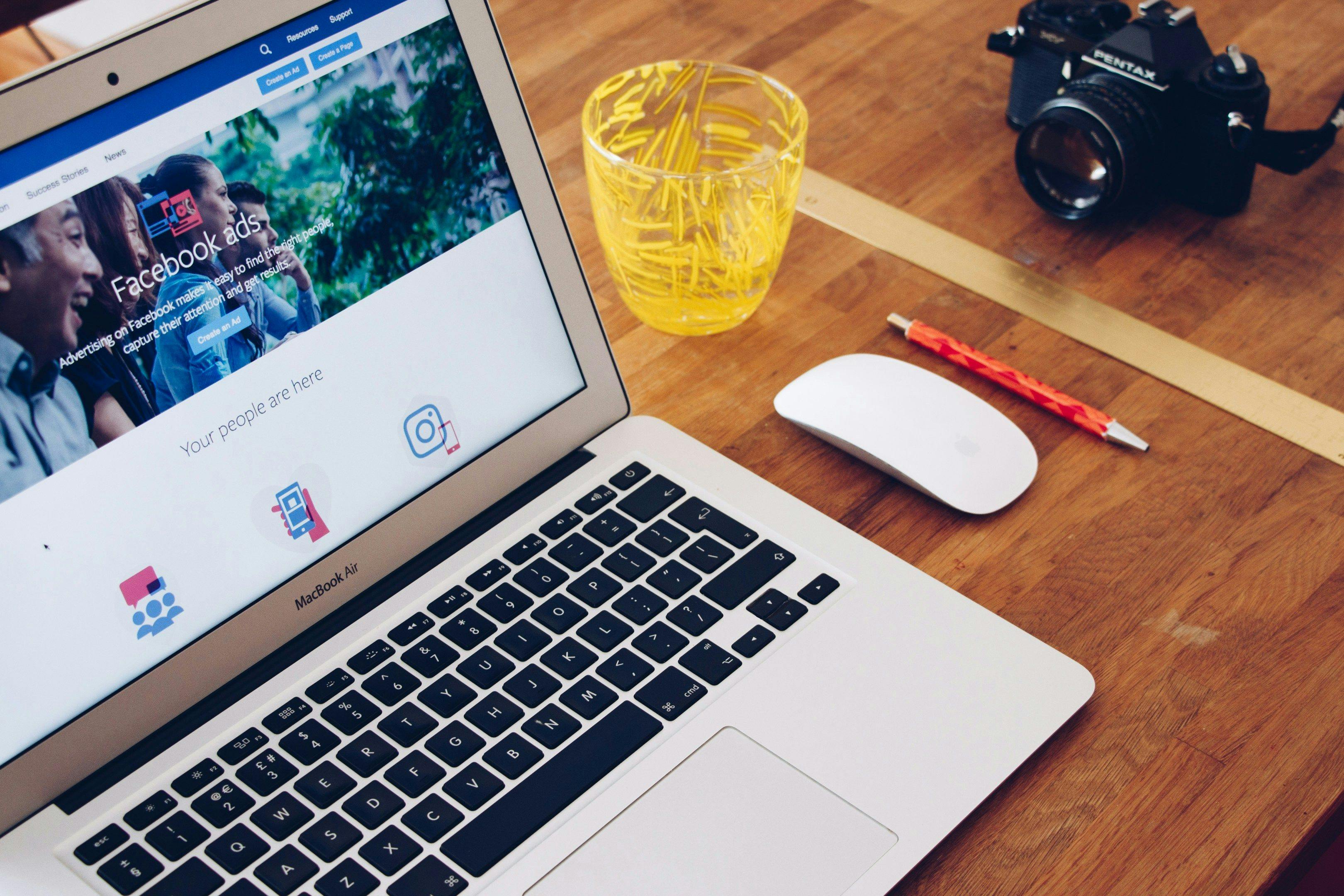 A laptop screen displaying a Facebook Ads page, surrounded by a camera, pencil, and a glass filled with pens, indicating a creative and organized workspace. This setup represents the tools and environment a "small business marketing coach" might use to teach social media marketing strategies.