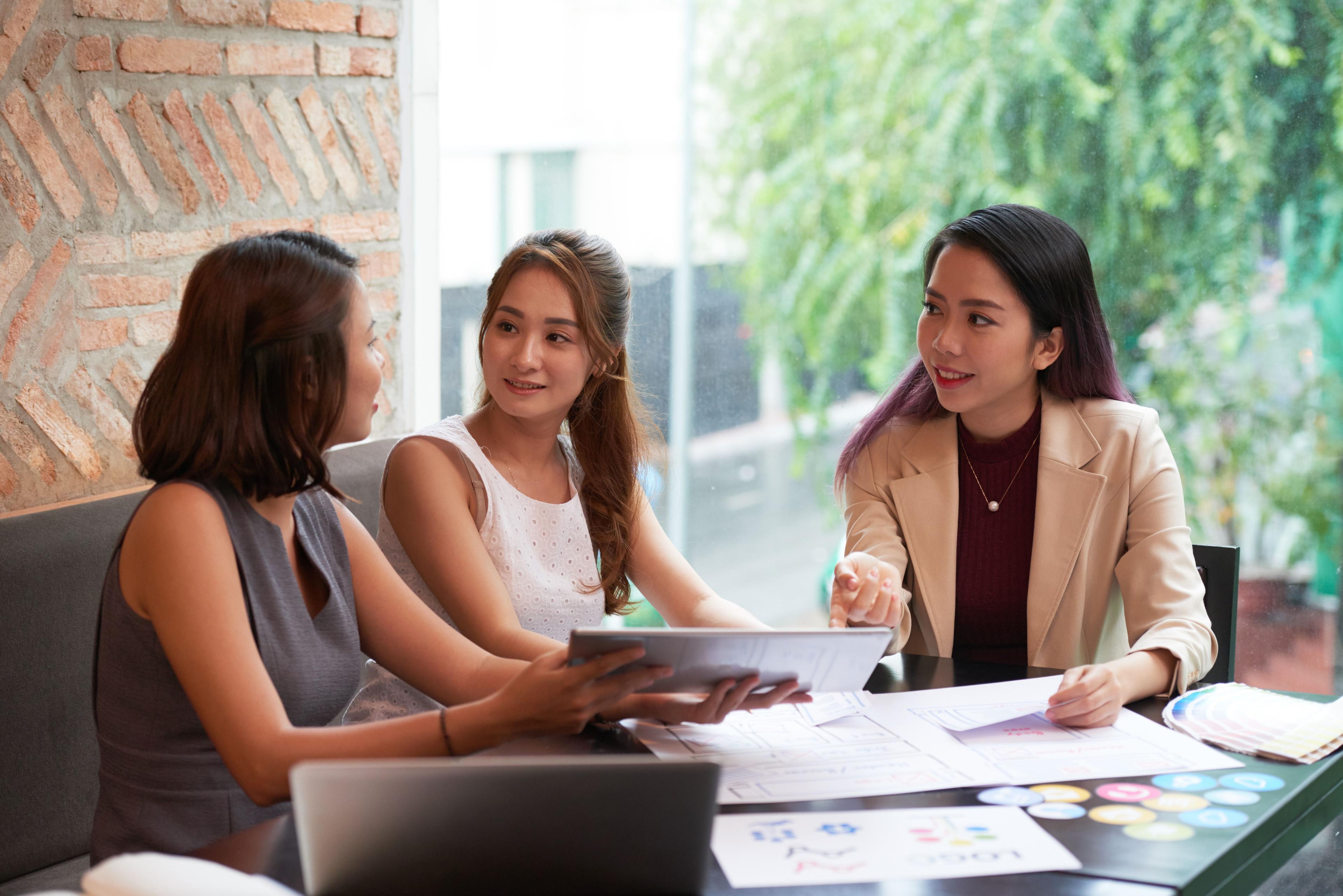 Three women are seated at a table with design materials, engaging in a lively discussion, representing a startup marketing agency planning a new campaign or branding strategy for a client.