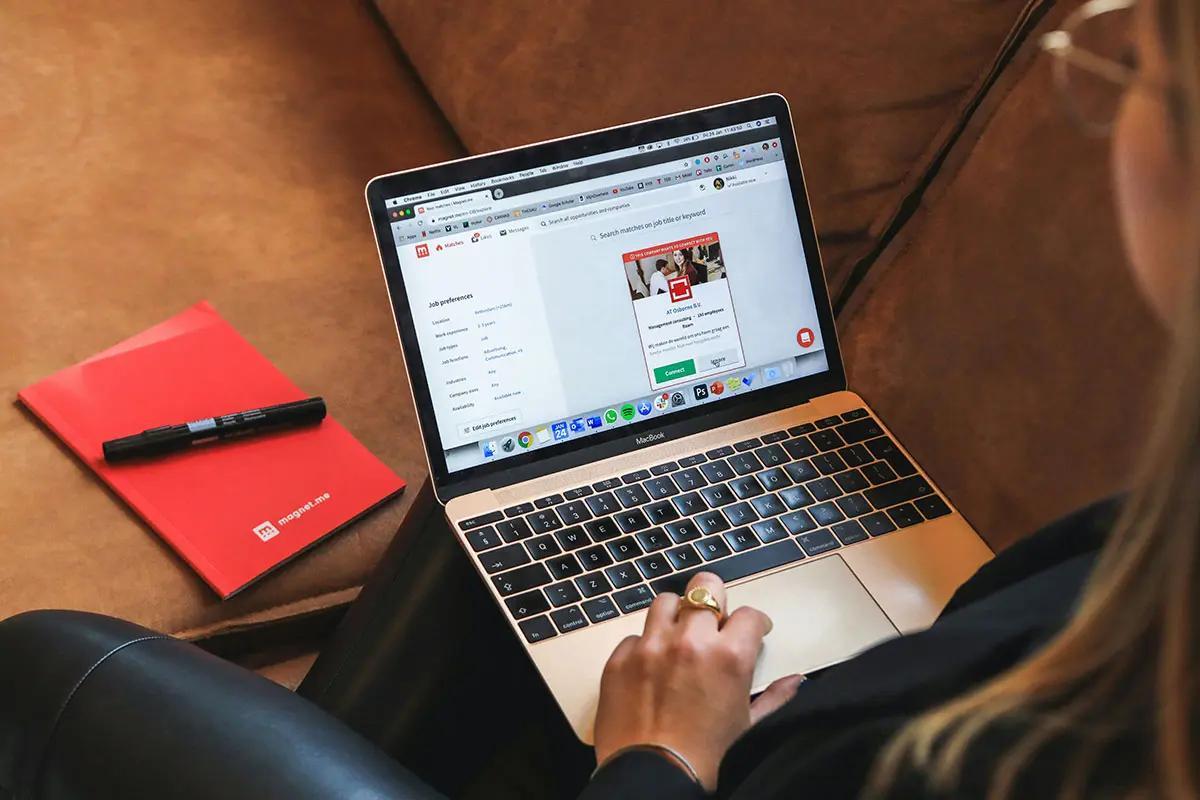 A person is sitting on a comfortable brown couch, using a laptop to browse a website that features job preferences. The laptop screen is visible, showing various tabs and a detailed webpage. Beside the person is a red notebook and a black marker. The setting is casual yet focused, making it suitable for illustrating the concept of "best AI image generator."