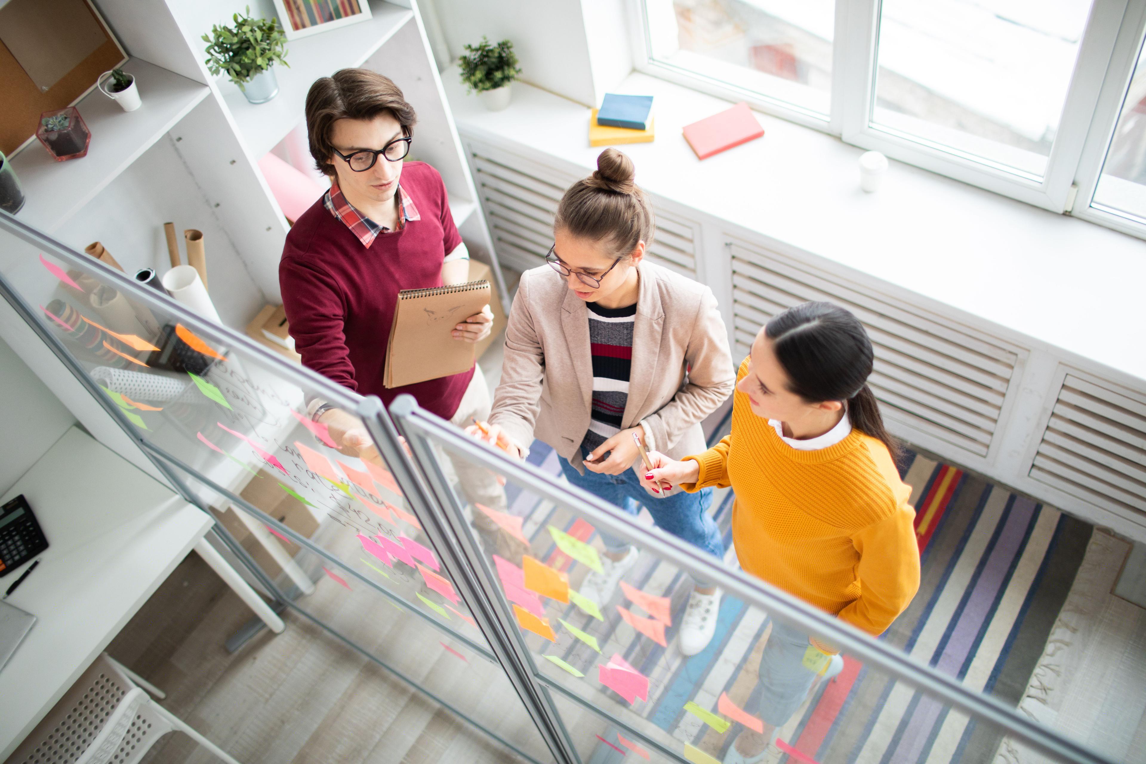 A team of three professionals brainstorming on stock market performance strategies, using sticky notes on a glass board in a contemporary workspace.