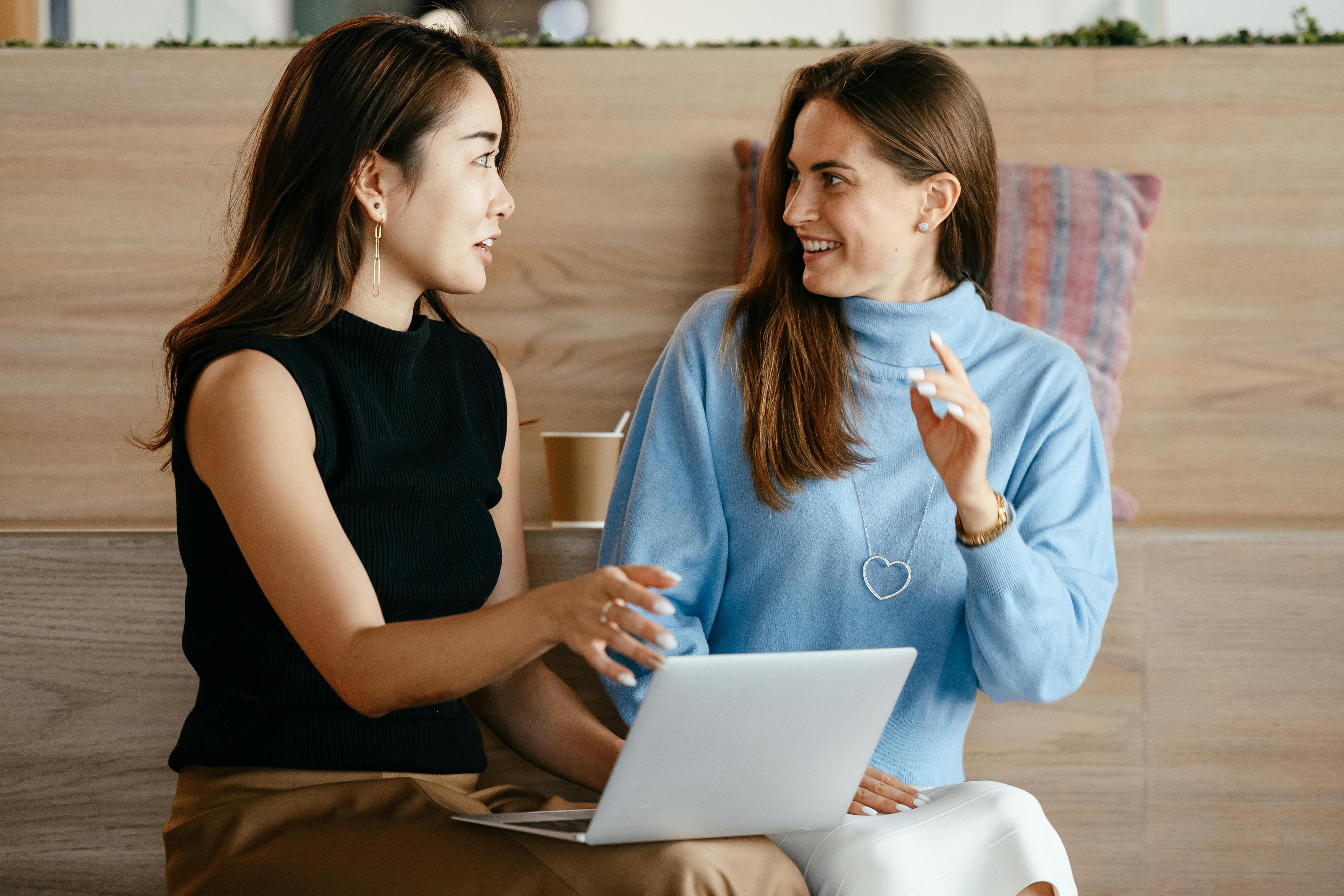 Two women are sitting and engaging in a lively discussion, one holding a laptop. They appear to be in a modern, comfortable setting. The image conveys collaboration and could be related to discussing AI in email marketing. A coffee cup and some pillows in the background add to the casual, yet professional atmosphere.