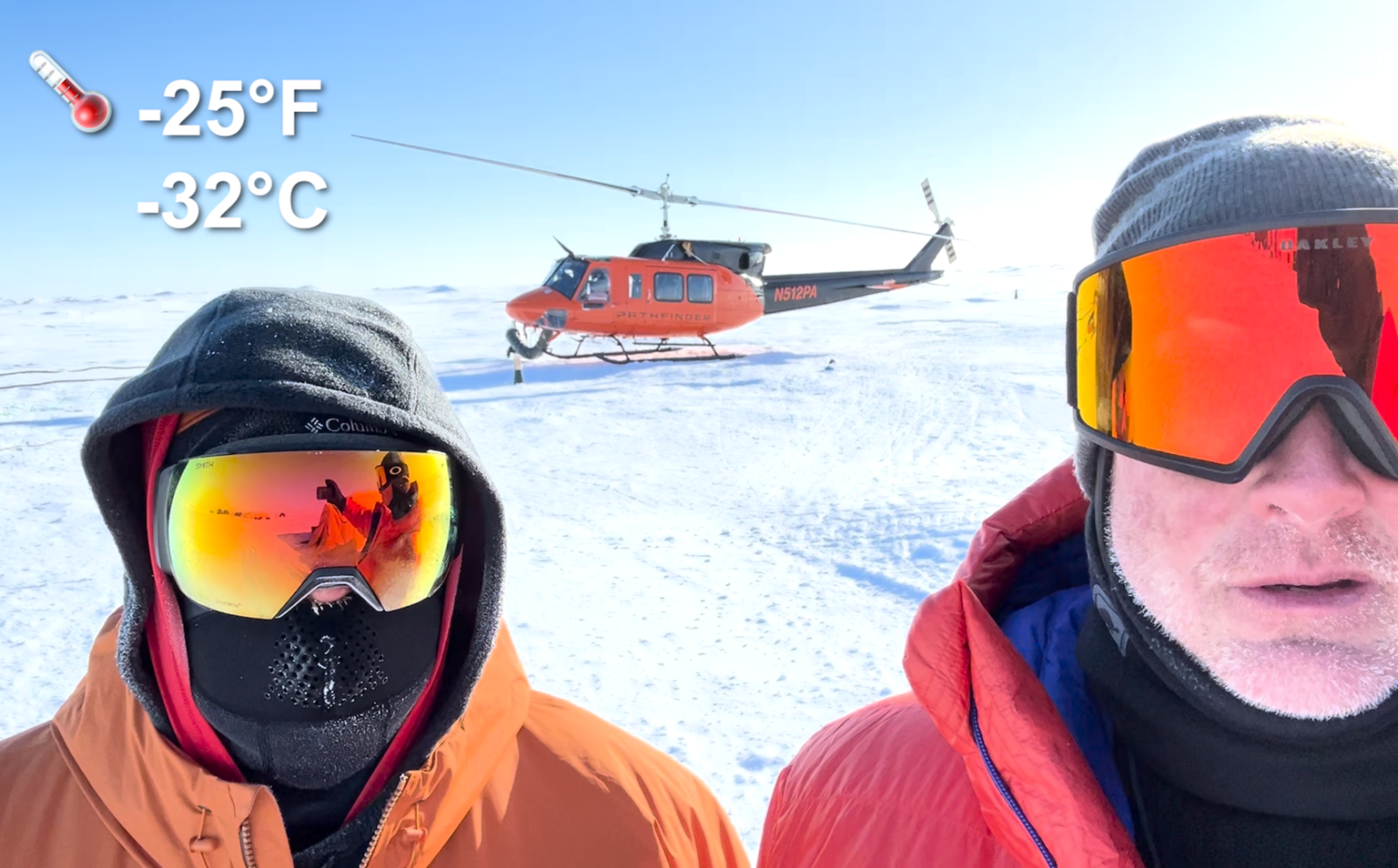 Ben Evans and Dave Whelihan stand bundled up outside in minus 25 degrees Fahrenheit in the Arctic, with a helicopter behind them, as they document their experience in the Arctic.