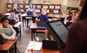 Image of a teacher checking her tablet in front of a classroom full of children at desks.