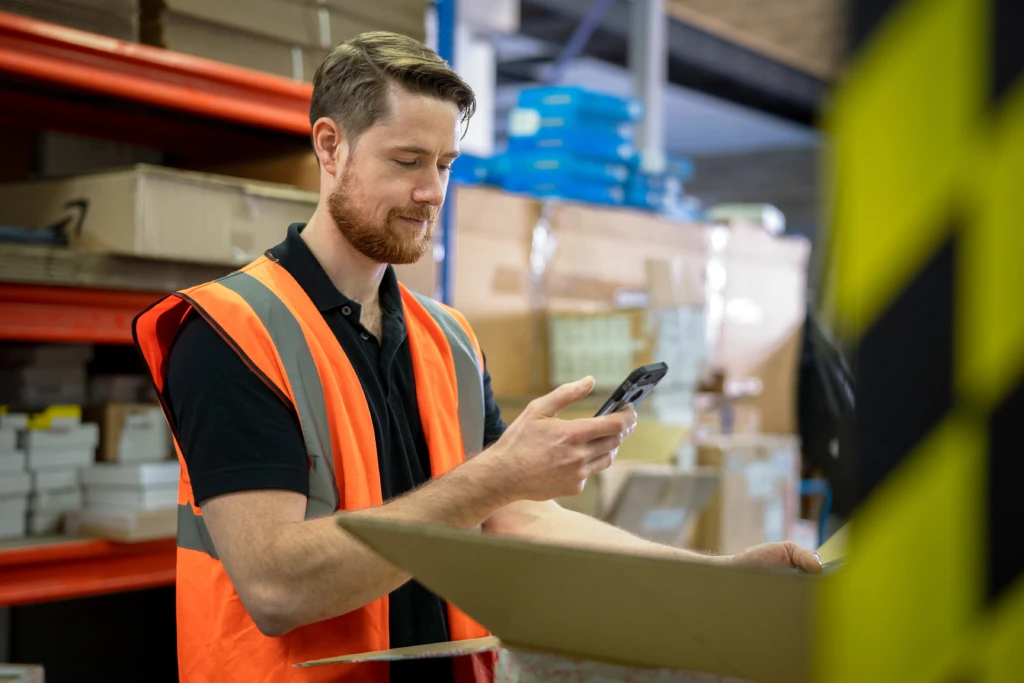 Man wearing an orange vest in a manufacturing or warehouse who appears to be doing inventory or looking at something in a box and messaging about it on his phone in Microsoft Teams. Key words: tele conferencing, cloud calling, Microsoft Teams, meeting, conference call, phone system, video system, Business Voice, Manufacturing, Firstline Worker, Delivery, Tracking