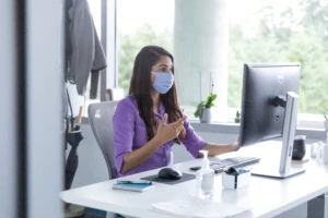 A female employee talking on a video call with hand sanitizer on her desk, socially distancing from others. Hybrid Workplace collection.