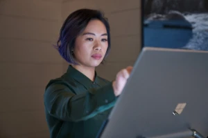 Side profile of a woman wearing a dark shirt in a dim office reaching up and working on a Microsoft Surface Studio. Keywords: touch screen, desktop, cloud security, threat protection, secure score, monitoring, Microsoft Security collection