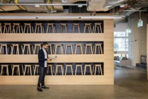 Male small business worker standing in front of three large shelves filled with dozens of wood stools. He is using an HP convertible laptop (opened as tablet) and pen. Screen is partially shown depicting bar charts and graphs.