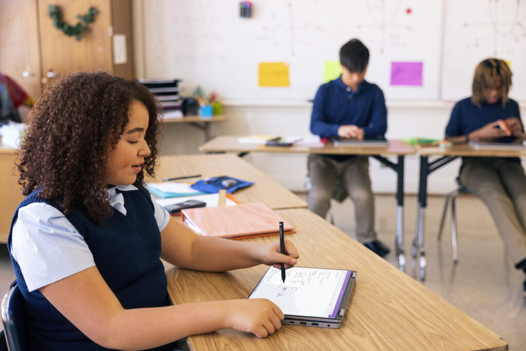 A student in a classroom writing on a laptop with a digital pen, with two other students working independently in the background.