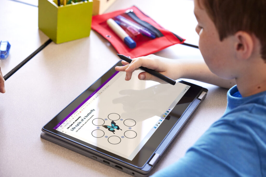 A student sitting at a desk in a classroom and using a digital pen to do an assignment on a laptop.