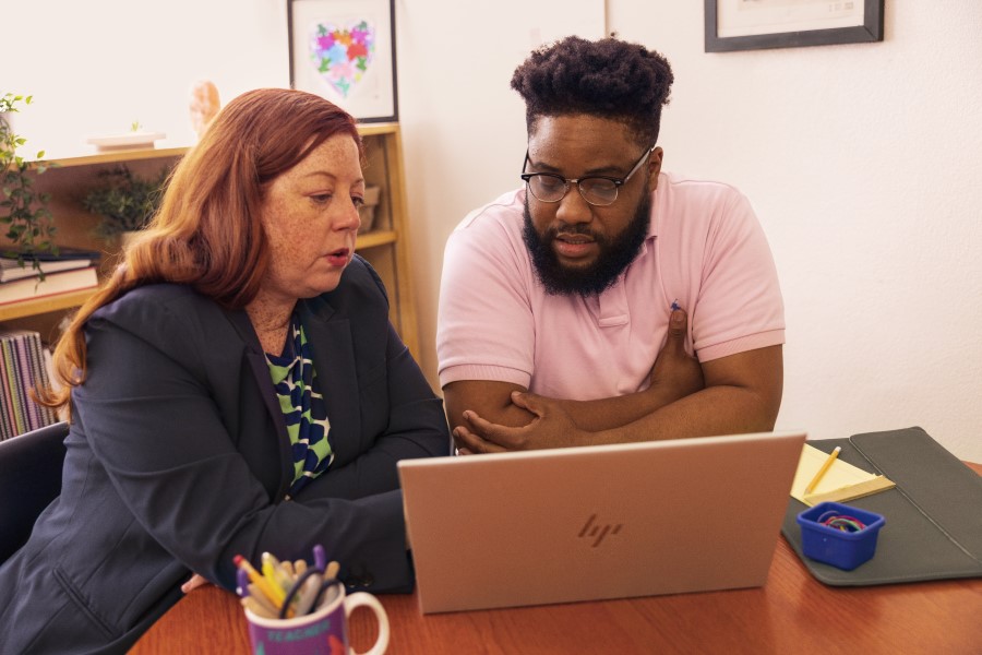 An educator and a school technologist sitting at a desk in a school office and looking at the screen of a laptop together.