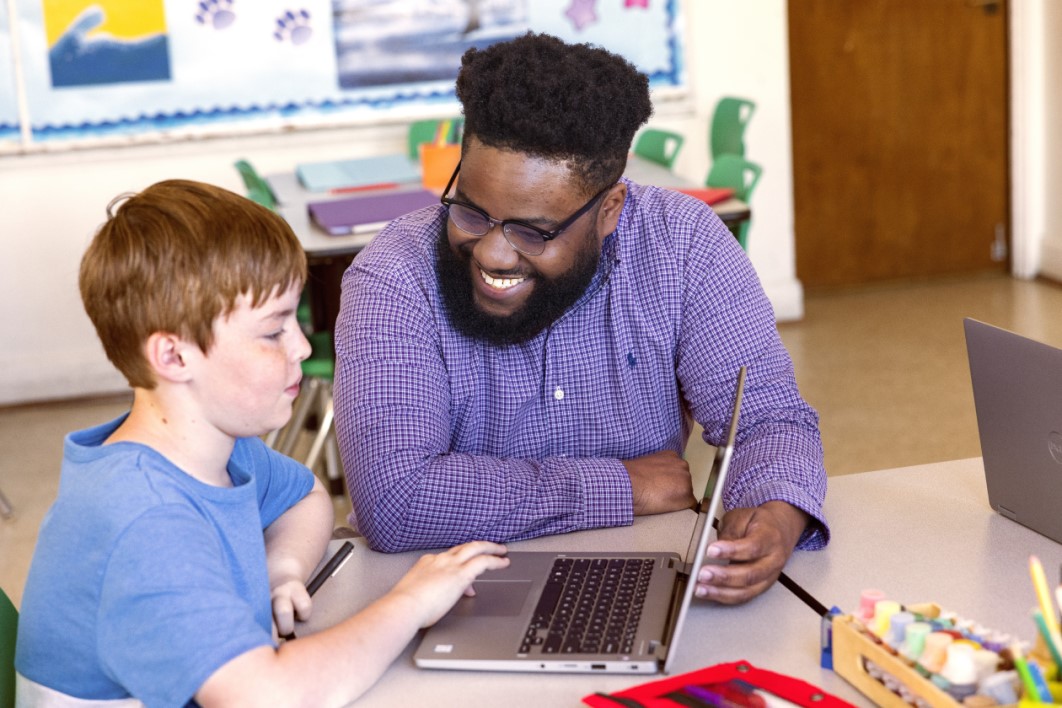 A teacher kneeling at a table engaging with a student who’s working on a laptop.