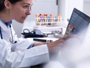 Person in laboratory with testing tubes of liquid next to them, looking at a computer screen.