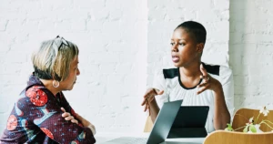 Two businesswomen sit at a table talking with a laptop in front of them.