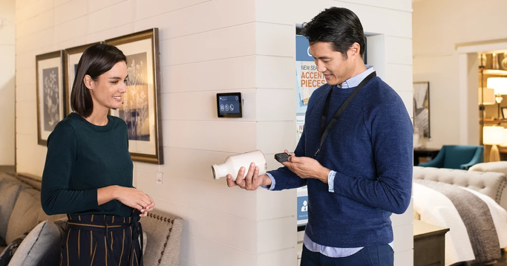 Male and female employees standing on home furnishings sales floor in commercial retail store. He is using Askey device to scan price tag on bottom of vase, while she looks on. A wall tablet is mounted nearby. Internet of things photography.