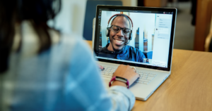 Image of a woman at a desk using a Surface laptop to make a Microsoft Teams video call with one man smiling and wearing a headset.