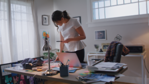 image of a woman standing over a desk at home, looking down at her laptop