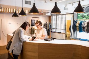 Female employee holding tablet for female consumer to sign and approve purchase. They are standing on either side of a retail clothing store checkout counter.