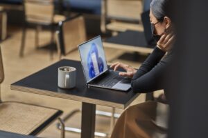 Woman with mask on for COVID compliance sitting in work space working on laptop with Windows 11 start screen image.