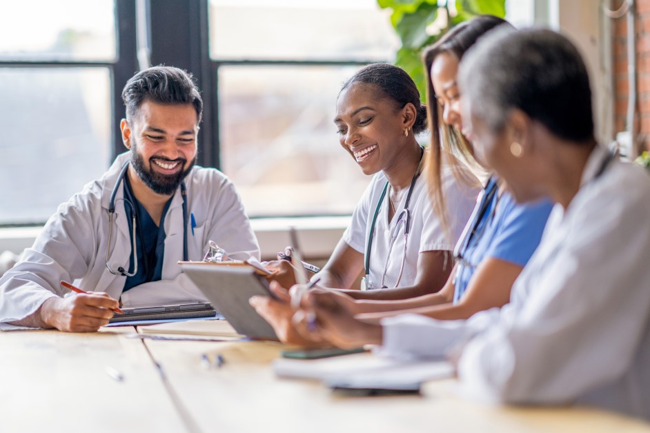 A small group of four medical professionals sit around a conference table as they meet to discuss patient cases. They are each dressed professionally in scrubs and lab coats as they focus on working together.