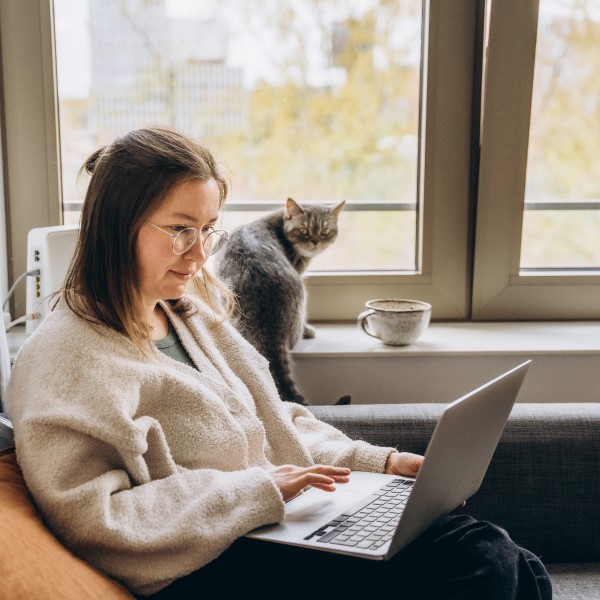 Young woman working from home uses a laptop while sitting on the sofa with her cat.