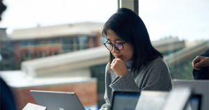 An image of a woman with glasses sitting at a community table in common area reading on a Surface laptop.