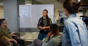 An image showing a female developer speaking in front of a white board during team stand up meeting.