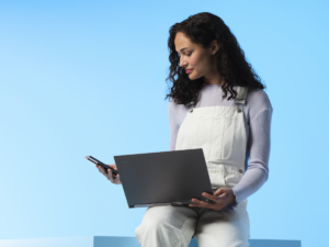 Photograph of young adult female using Lenovo YOGA SLIM 7 and mobile phone in studio on blue background.