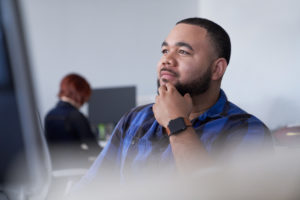 Man with beard sitting behind a computer monitor with his hand to his chin thinking or looking up.