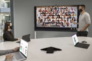 Two adults in masks collaborating while on a Microsoft Teams Together Mode meeting on a Surface Hub 2S 85” device.