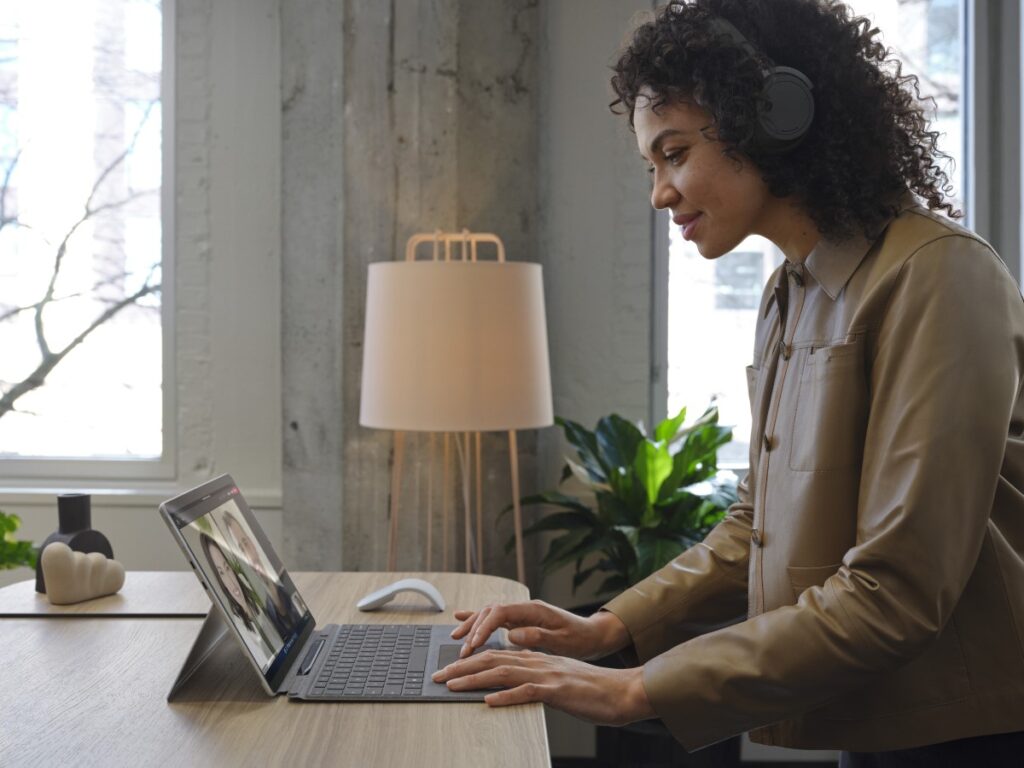 Adult female wearing Surface Headphones 2+ standing at a desk, working on a Surface Pro X in Microsoft Outlook.