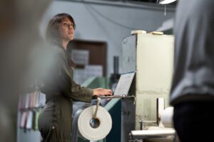 CEO of textile manufacturing plant working on shop floor analyzing data on her laptop.