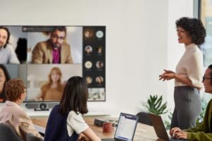 A female speaker presenting in a Microsoft Teams Room meeting while people in the conference room are viewing remote attendees in Gallery Mode with participant chat on the front of room screen​.