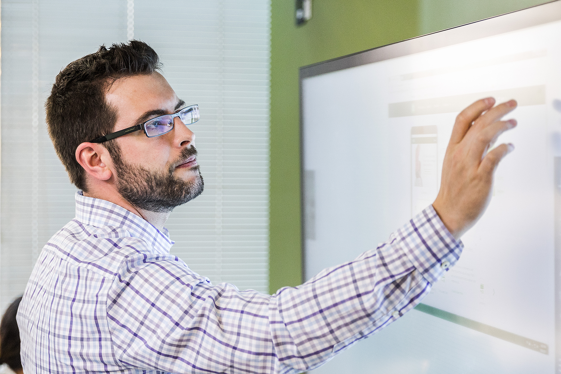 Male office worker standing in modern office and using large touchscreen monitor with right hand (screen shows open browser window).