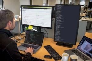 Photo of Male developer is coding at his workspace in an enterprise office using Visual Studio on a MacBook Pro multi-monitor set up.