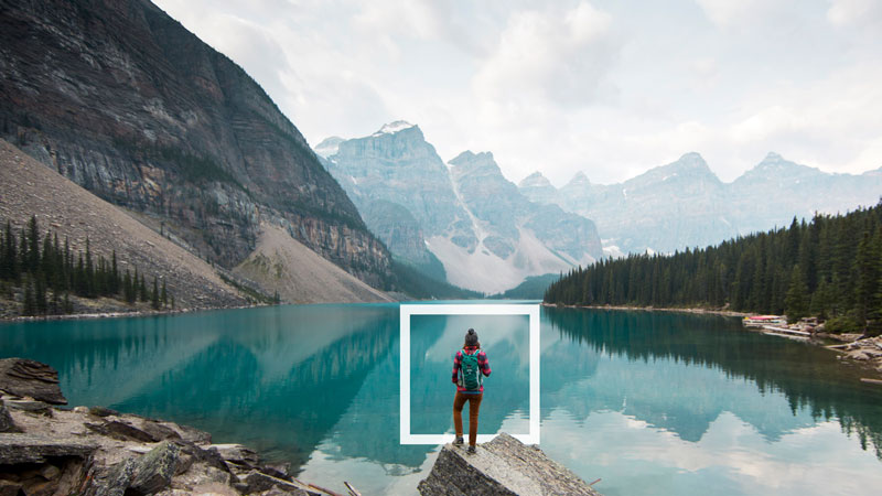 A person stands on a rock overlooking a glacier lake with a forest and mountains in the distance