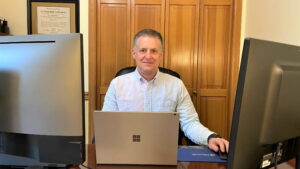 Pete Fortman is sitting at a desk in his home office in a blue-striped collared shirt. A laptop and two monitors with no information displayed are visible.