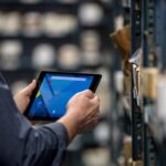 Male employee holding a tablet with both hands. He is standing in front of a metal warehouse rack filled with packages.