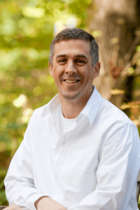 Bogdan smiles with his arms crossed in front of him in a photo taken in front of greenery outdoors.