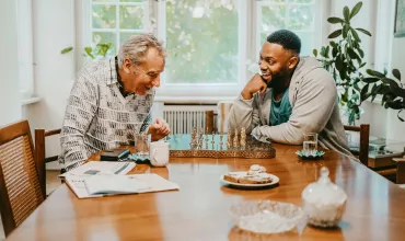 Two men, seated indoors around a table, engaged in a conversation and playing chess