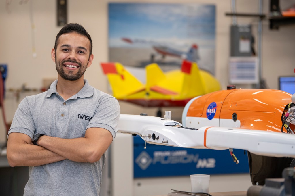 A man wearing a gray NASA shirt posing to the left of a subscale model or an aircraft that is orange and white with the NASA meatball on it. In the background, there are aviation posters located on the wall and another subscale model on display that is yellow and red.