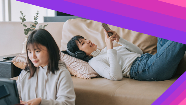 One women working over laptop and another women lying on sofa - listening music via phone