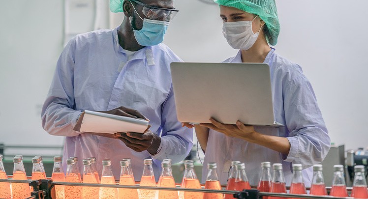 Two people in protective suits working on a laptop in a factory.