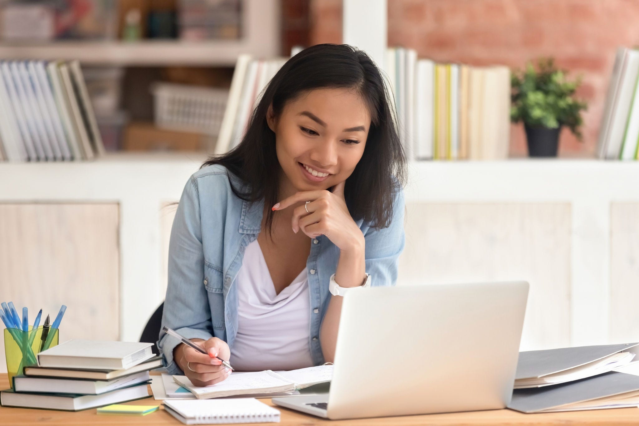 Smiling asian student studying in library with laptop books doing online research for coursework, making notes for essay homework assignment, online education e-learning concept