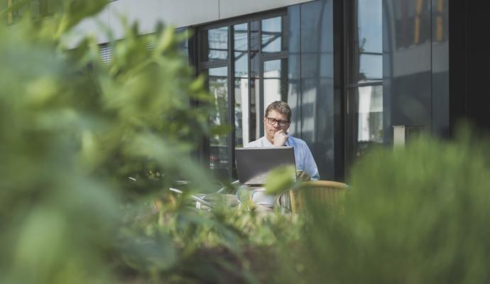 Photo of a man sitting outside with a laptop, with shrubbery in the foreground 