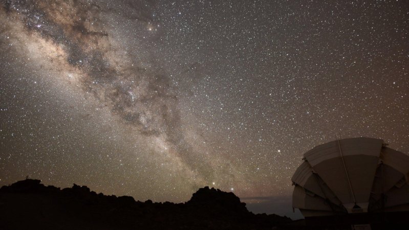 The Milky Way galaxy shines above a dark space telescope in the foreground.