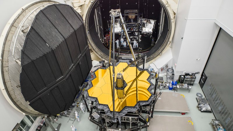 The gold mirrors of JSWT sit in front of an extremely large, circular door in a vast room with scientists working.