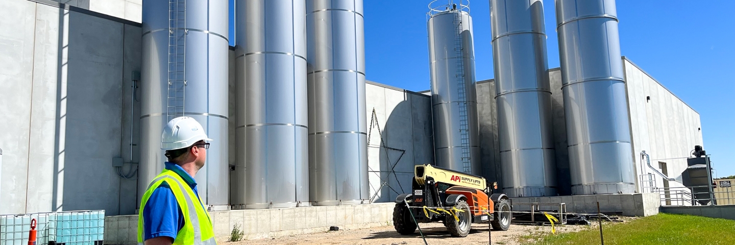 A man in a hard hat and safety vest looks at six tall metal tanks next to a building. 